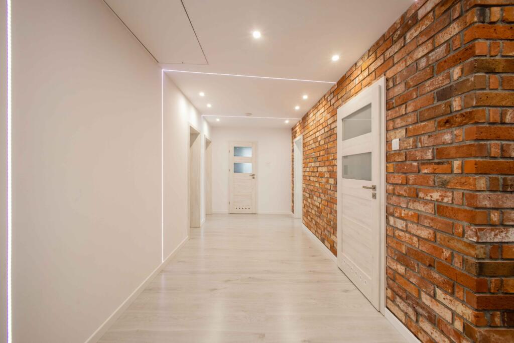 Nowoczesne korytarz w którym znajdują się białe ściany i biały sufit wyposażony w paski ledowe, po prawej stronie znajduje się ceglana ściana wykonana z płytek z naturalnej cegły