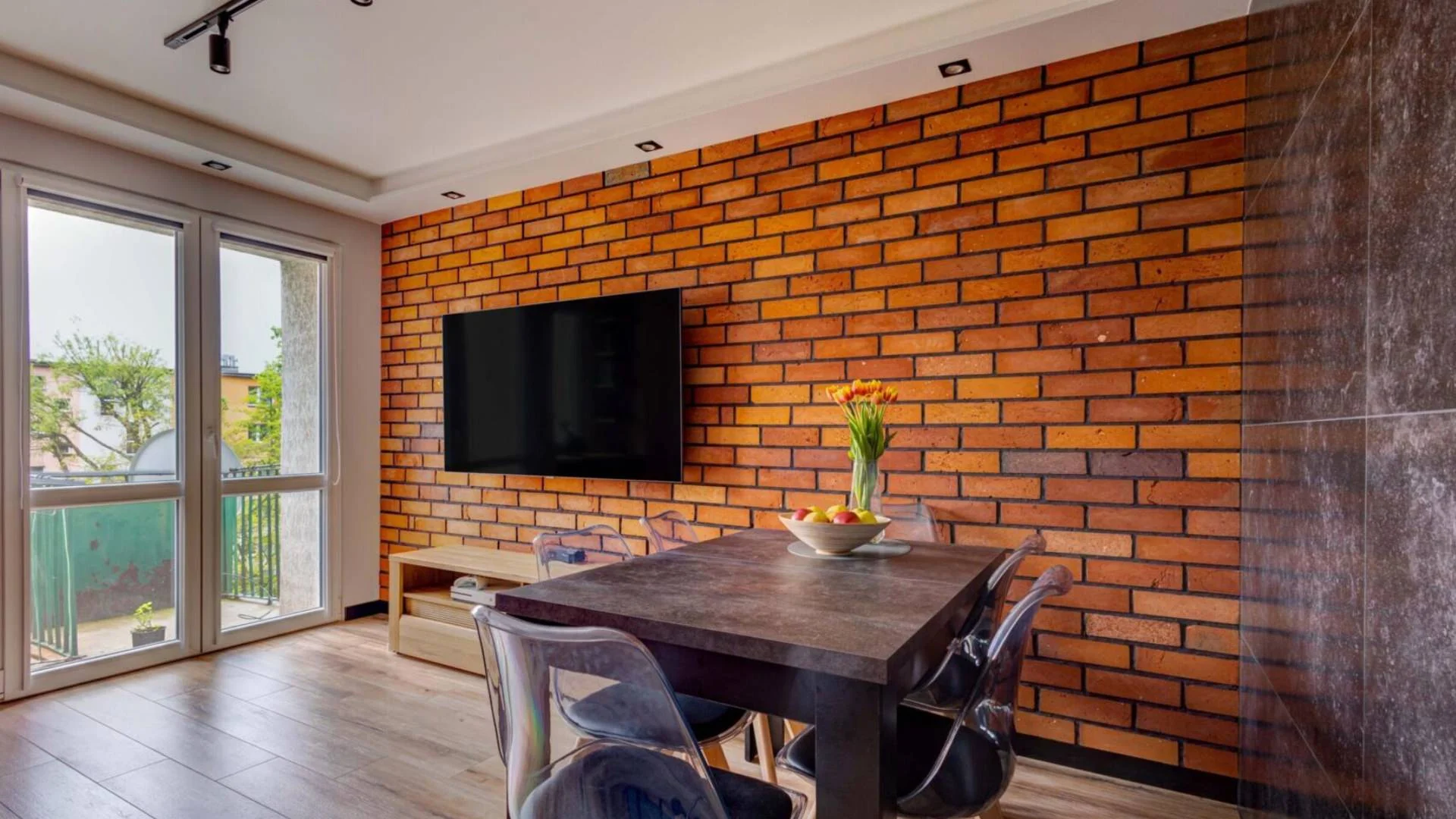 Nowoczesny salon z jadalnią, w którym ceglana ściana nadaje wnętrzu ciepła i charakteru. Minimalistyczny stół z ciemnego drewna i transparentne krzesła dodają elegancji.