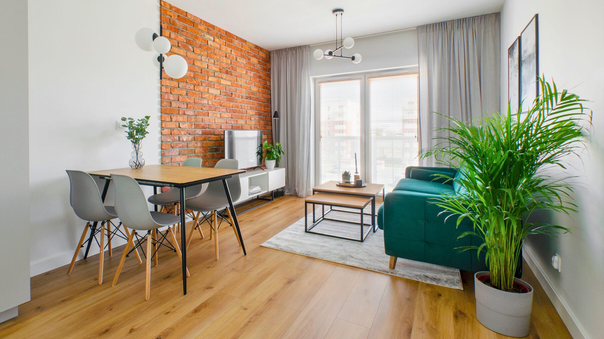 Nowoczesny salon z czerwono-ceglaną ścianą, drewnianą podłogą i minimalistycznym wystrojem. Zielona sofa oraz rośliny wprowadzają naturalny, przytulny klimat.