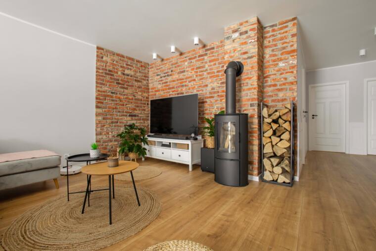 Salon wyposażony w ścianę telewizyjną z cegły oraz drewnianą podłogę, stelaż na drewno kominkowe oraz kominek