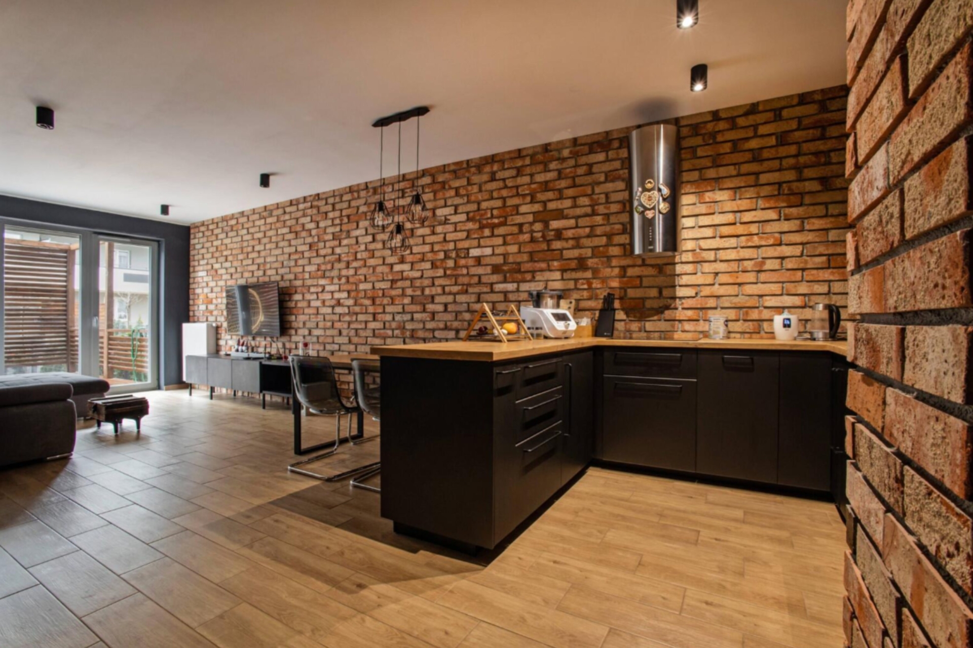 Nowoczesny salon z kuchnią i ścianą z cegły rozbiórkowej, czarne meble i drewniana podłoga